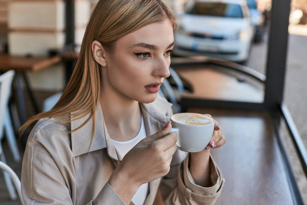 dromerige jonge vrouw in trench jas genieten van haar kopje cappuccino terwijl zitten buiten in cafe - Foto, afbeelding