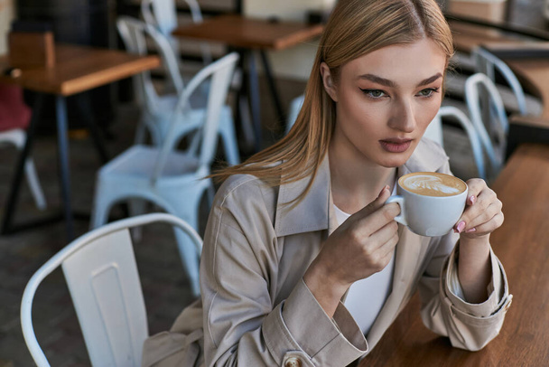 мечтательная красивая женщина в плаще наслаждается чашкой капучино, сидя на улице в кафе - Фото, изображение