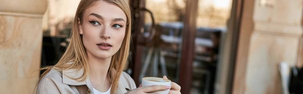dreamy and young woman in trench coat enjoying her cup of cappuccino while sitting in cafe, banner - Photo, Image