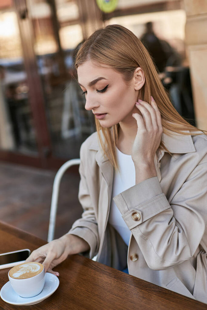 attractive woman in trench coat enjoying cup of cappuccino while sitting near smartphone in cafe - Foto, Bild