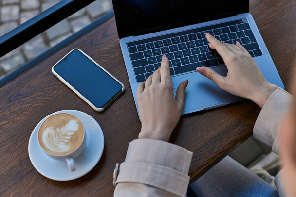 вид женщины, печатающей на ноутбуке, работающей дистанционно возле чашки кофе и смартфона в кафе - Фото, изображение