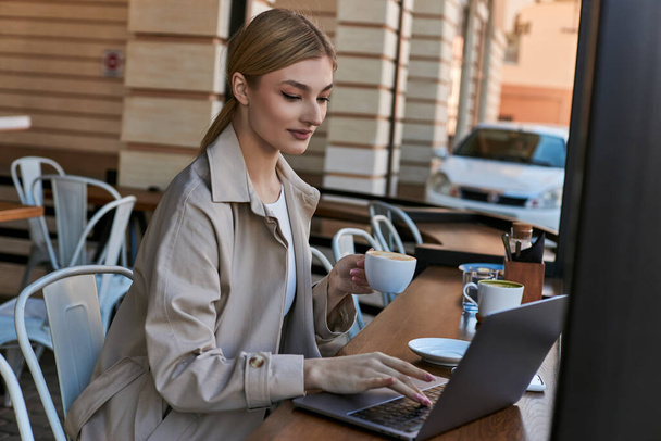 jeune femme blonde tenant une tasse de café et utilisant un ordinateur portable sur la table dans un café à l'extérieur, travail à distance - Photo, image