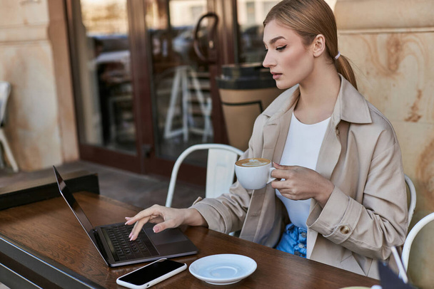 blondi nainen tilalla kuppi kahvia ja käyttää kannettavaa pöydällä kahvilassa ulkona, etätyö - Valokuva, kuva