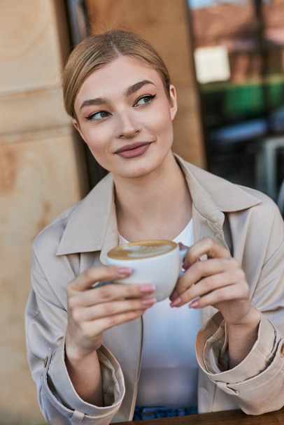 mooie blonde vrouw in trench jas genieten van haar kopje cappuccino in cafe, ontspannen sfeer - Foto, afbeelding