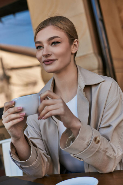блондинка в плаще наслаждается чашкой капучино в кафе, расслабляющая атмосфера - Фото, изображение