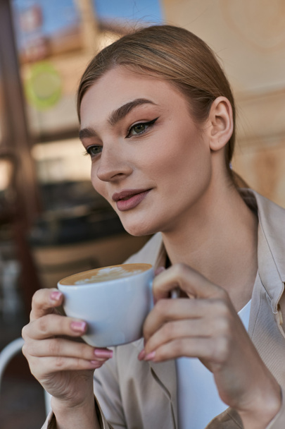 элегантная блондинка в плаще наслаждается чашкой капучино в кафе, расслабляющая атмосфера - Фото, изображение