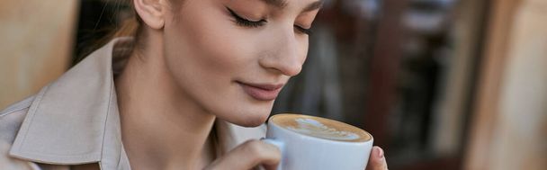 банер красивої блондинки в тренч пальто насолоджуючись чашкою капучино в кафе - Фото, зображення