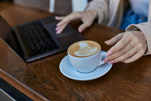 abgeschnittene Aufnahme einer jungen Frau im Trenchcoat mit Laptop in der Nähe einer Tasse Cappuccino im Café, Fernarbeit - Foto, Bild