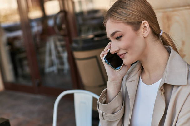Sonbahar bej renkli trençkotlu mutlu genç kadın kafede gülümsüyor ve akıllı telefonuyla konuşuyor. - Fotoğraf, Görsel