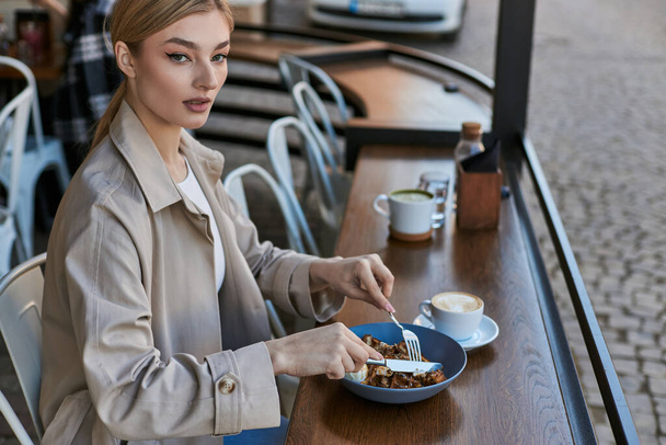 mooie jonge vrouw in trench jas eten haar belgische wafels met ijs naast kopje koffie - Foto, afbeelding