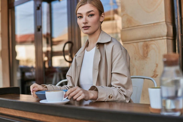 mooie jonge vrouw in stijlvolle trench jas zitten naast bord en kopje koffie in modern cafe - Foto, afbeelding