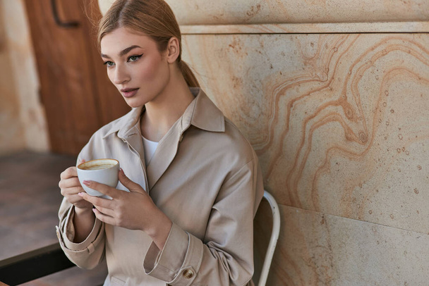 blonde young woman in stylish trench coat sitting and holding cup of coffee inside of modern cafe - Photo, Image