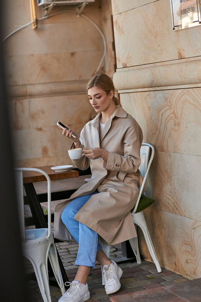 attractive young woman in stylish trench coat using smartphone and holding cup of coffee in cafe - Photo, Image