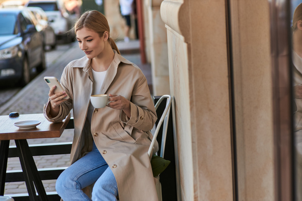 atrakcyjna kobieta w stylowym płaszczu Trench za pomocą smartfona w pobliżu filiżanki kawy na stole w kawiarni - Zdjęcie, obraz