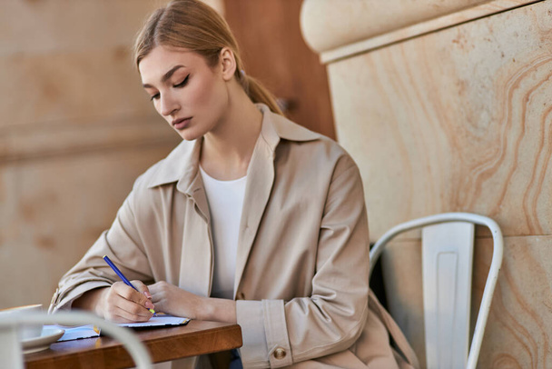 pretty young woman in stylish trench coat writing in notebook near cup of coffee, diary - Photo, Image