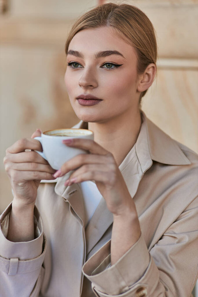 ładna blondynka kobieta w stylowy płaszcz Trench trzymając filiżankę kawy podczas siedzenia w kawiarni, napój - Zdjęcie, obraz