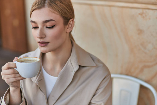 elegant blonde woman in stylish trench coat holding cup of coffee while sitting in cafe, cappuccino - Photo, Image