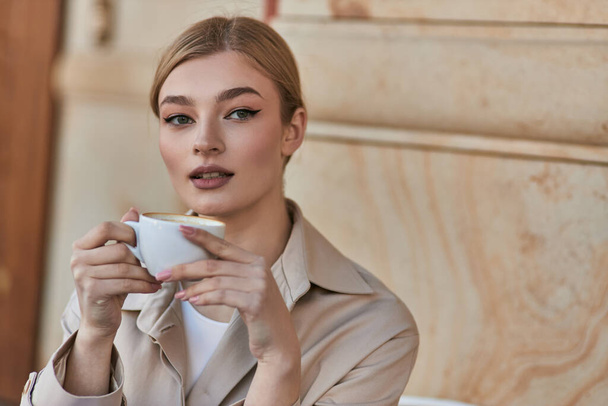 elegante giovane donna in elegante trench in possesso di una tazza di caffè mentre seduto in caffè, bevanda - Foto, immagini