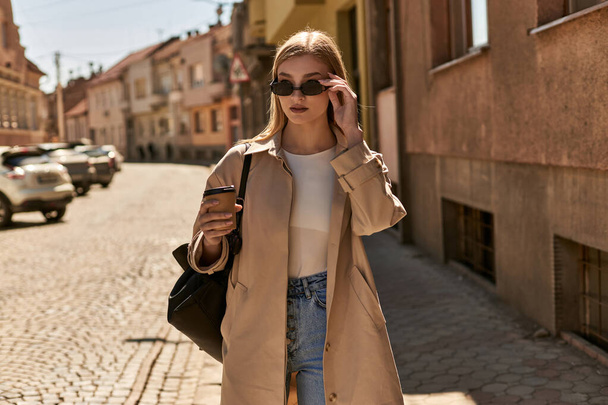mladá blondýna žena v trenč kabát držení odnést kávu, zatímco stojí na ulici ve slunečný den - Fotografie, Obrázek