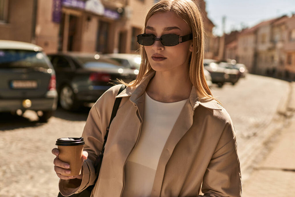スタイリッシュなトレンチコートのブロンド女性は,晴れた日の通りに立って行くためにコーヒーを保持しています - 写真・画像