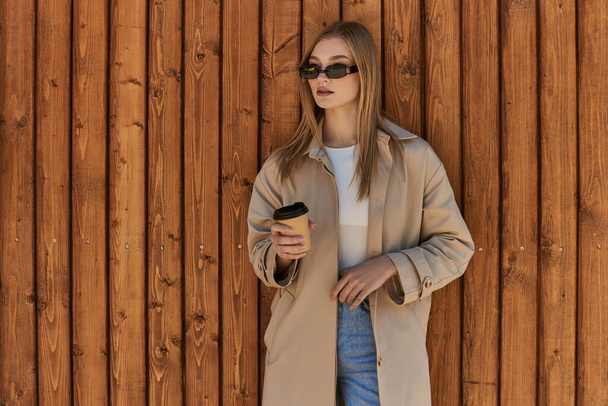 blondynka młoda kobieta w płaszczu i okulary przeciwsłoneczne trzymając kawę iść i stojąc w pobliżu drewnianego ogrodzenia - Zdjęcie, obraz