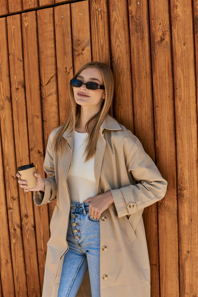 szczęśliwa kobieta w płaszczu i okularach przeciwsłonecznych trzymając kawę na wynos i stojąc z ręką w kieszeni - Zdjęcie, obraz