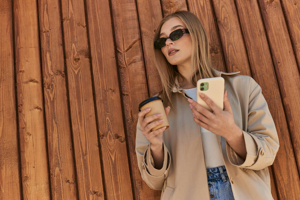 フェンス近くのスマートフォンを使用して行くためにコーヒーを保持するトレンチコートとサングラスのスタイリッシュな女性 - 写真・画像