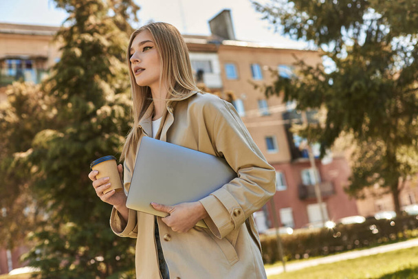 élégante femme blonde en trench coat marche avec ordinateur portable et café à emporter, pigiste en ville - Photo, image
