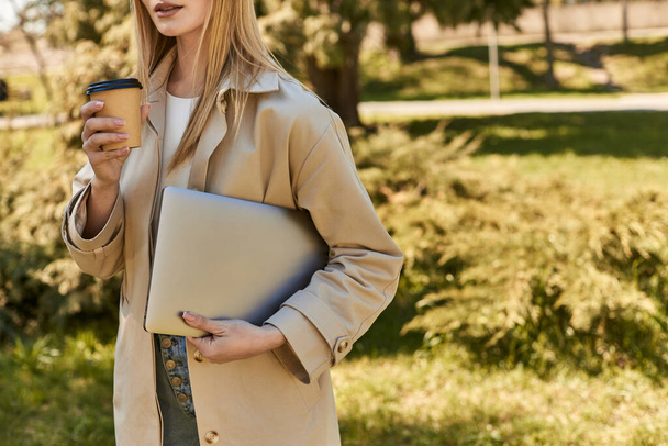 cropped young woman in trench coat holding laptop and drinking takeaway coffee, freelancer in city - Photo, Image