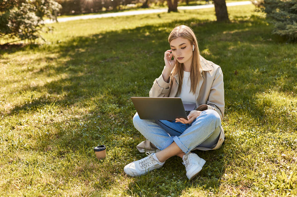 young blonde woman in earphones and trench coat sitting on grass near paper cup and using laptop - Photo, Image