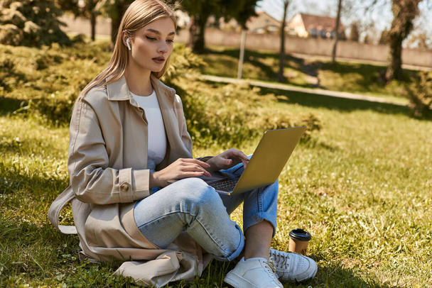 szőke fiatal nő fülhallgató és ballonkabát segítségével laptop, miközben ül a füvön mellett papír pohár - Fotó, kép