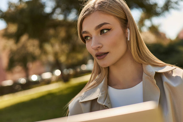 portrait of pretty young woman in wireless earphones and beige trench coat looking away in park - Photo, Image