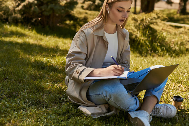 mulher loira em fones de ouvido e casaco de trincheira fazendo pesquisa on-line no laptop enquanto escreve no notebook - Foto, Imagem