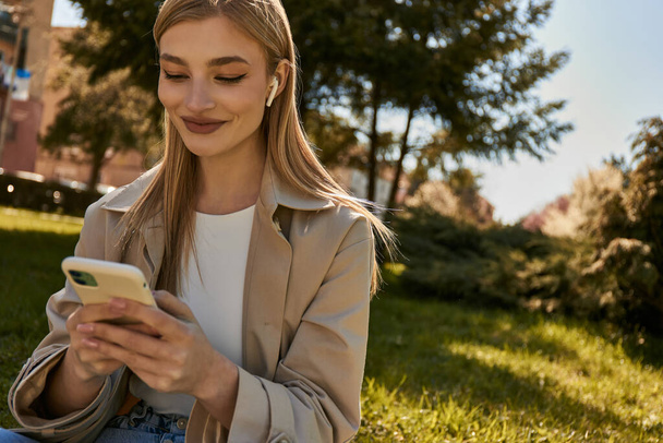 szczęśliwa blondynka w słuchawki bezprzewodowe i stylowy płaszcz Trench za pomocą smartfona w parku - Zdjęcie, obraz