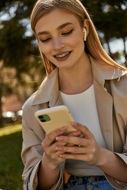 szczęśliwa blondynka w bezprzewodowych słuchawkach i beżowy płaszcz Trench za pomocą smartfona w parku - Zdjęcie, obraz