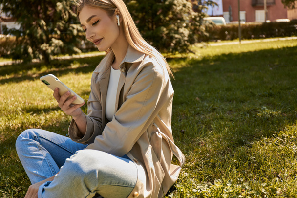 happy blonde woman in wireless earphones and trench coat using her smartphone and sitting on lawn - Photo, Image
