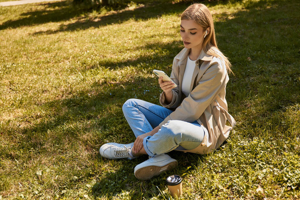 young blonde woman in wireless earphones and trench coat using her smartphone and sitting on lawn - Foto, afbeelding