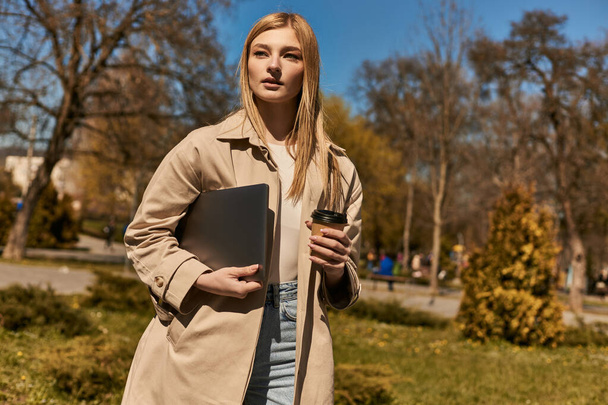 pretty blonde woman in stylish trench coat holding paper cup with coffee and laptop in park - Photo, Image
