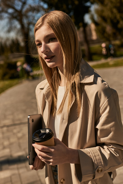 ベージュトレンチコートの若いブロンド女性コーヒーとラップトップとパークで歩く紙コップを保持 - 写真・画像