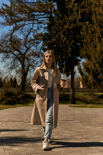 young blonde woman in trench coat holding paper cup with to go coffee and laptop and walking in park - Photo, Image