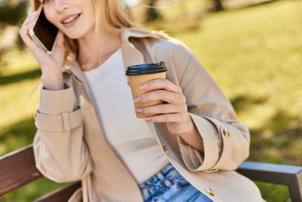 περικοπή γυναίκα σε καμπαρντίνα κρατώντας καφέ για να πάει και μιλώντας στο smartphone, ενώ κάθεται στον πάγκο - Φωτογραφία, εικόνα