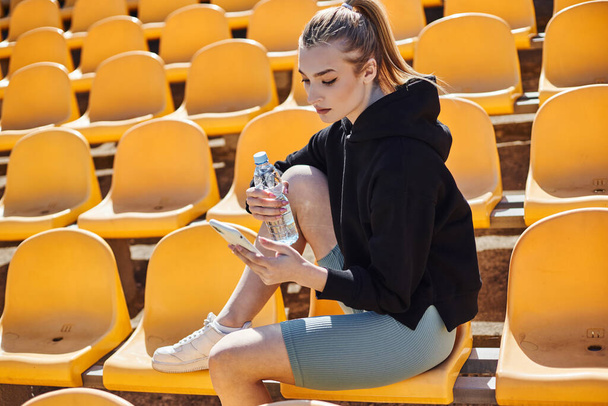 sportovní žena s ohonem drží láhev vody a pomocí smartphone po cvičení na stadionu - Fotografie, Obrázek