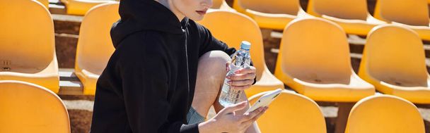 Kırpılmış sporcu kadın elinde bir şişe su tutuyor ve antrenmandan sonra stadyumda akıllı telefon kullanıyor. - Fotoğraf, Görsel