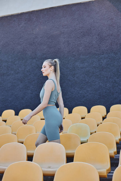 athletische und blonde Sportlerin in aktiver Kleidung, die durch gelbe Stadionstühle läuft, Sportmode - Foto, Bild