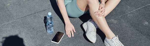 banner ritagliato di sportiva in activewear seduta accanto a bottiglia d'acqua e smartphone sul pavimento - Foto, immagini