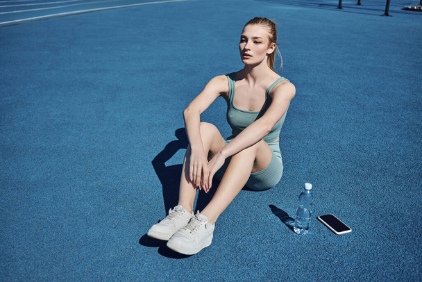 junge athletische Frau in Aktivkleidung sitzt nach dem Training neben Wasserflasche und Handy - Foto, Bild