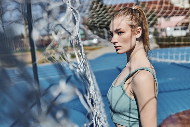 blonde and fit woman with ponytail standing in activewear near net after working out outdoors - Photo, Image