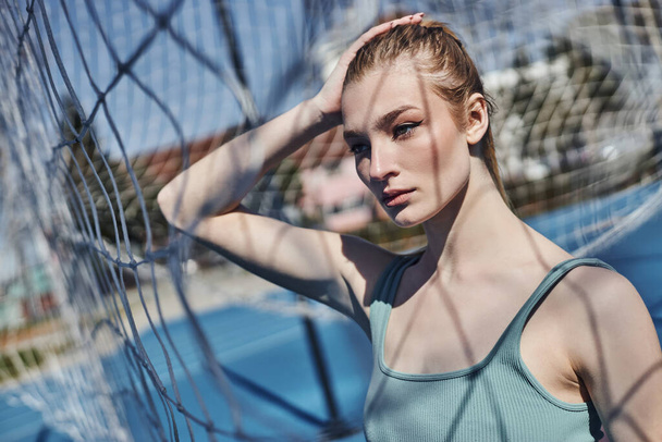 portrait of blonde and fit woman standing in activewear near net after working out outdoors - Photo, Image