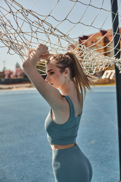 blondýny mladá žena s culíkem pózování v activewear při stání v blízkosti sítě po cvičení - Fotografie, Obrázek