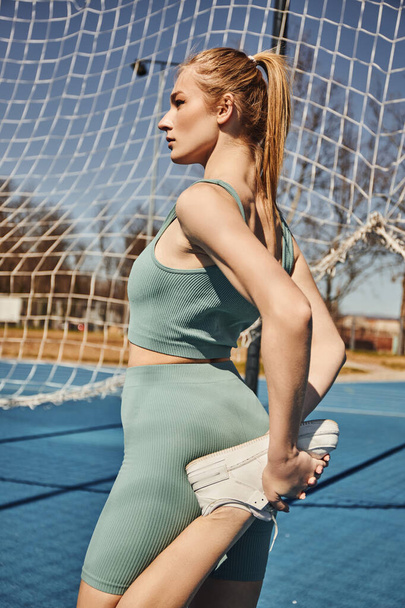 blonde young sportswoman with ponytail exercising in activewear near net outdoors, stretching leg - Photo, Image